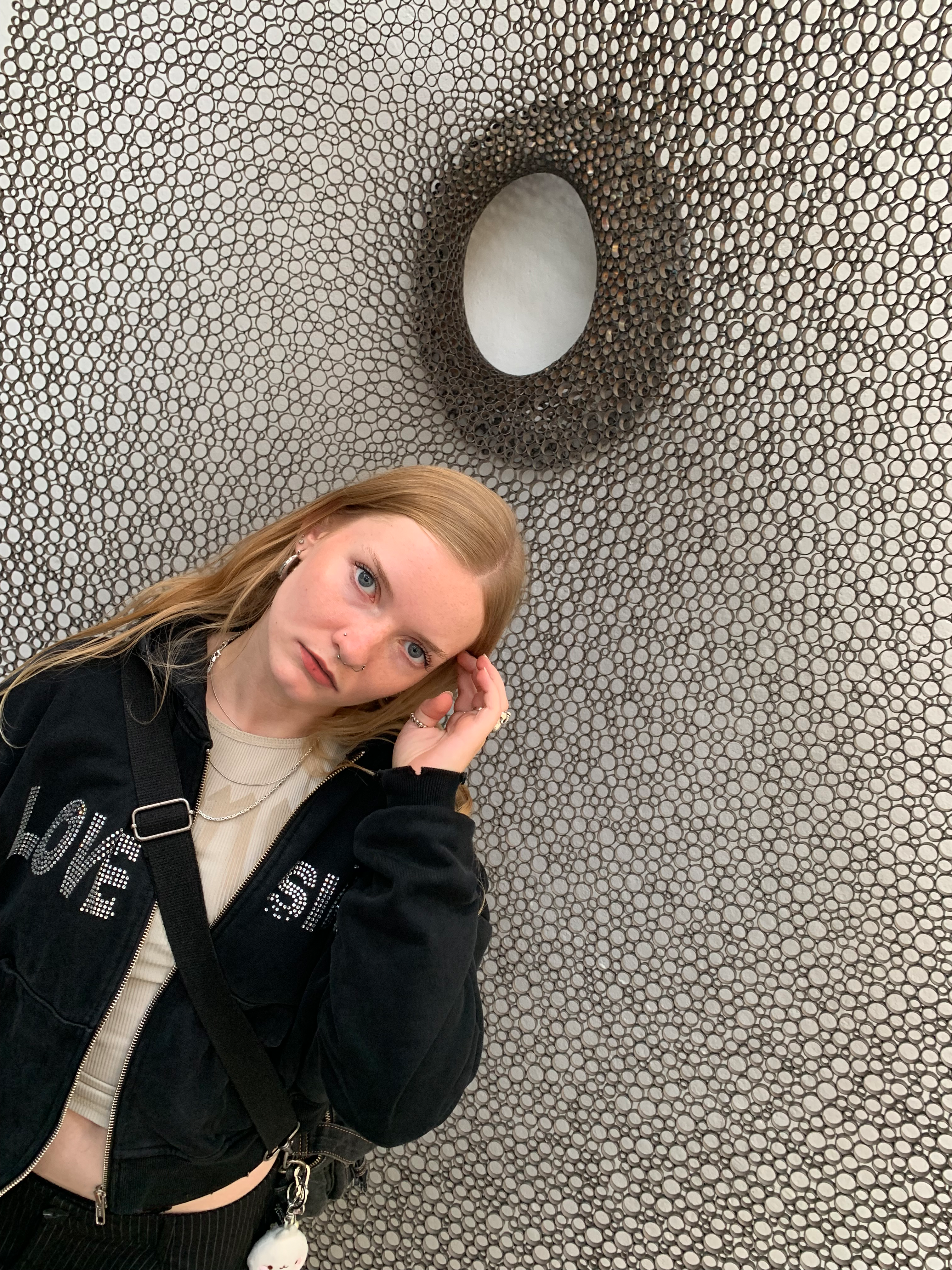 Black and white photograph of a young woman standing in front of a textured wall made of circular metallic elements. She wears a black zip hoodie and tilts her head slightly while looking at the camera, creating a relaxed and thoughtful mood.
