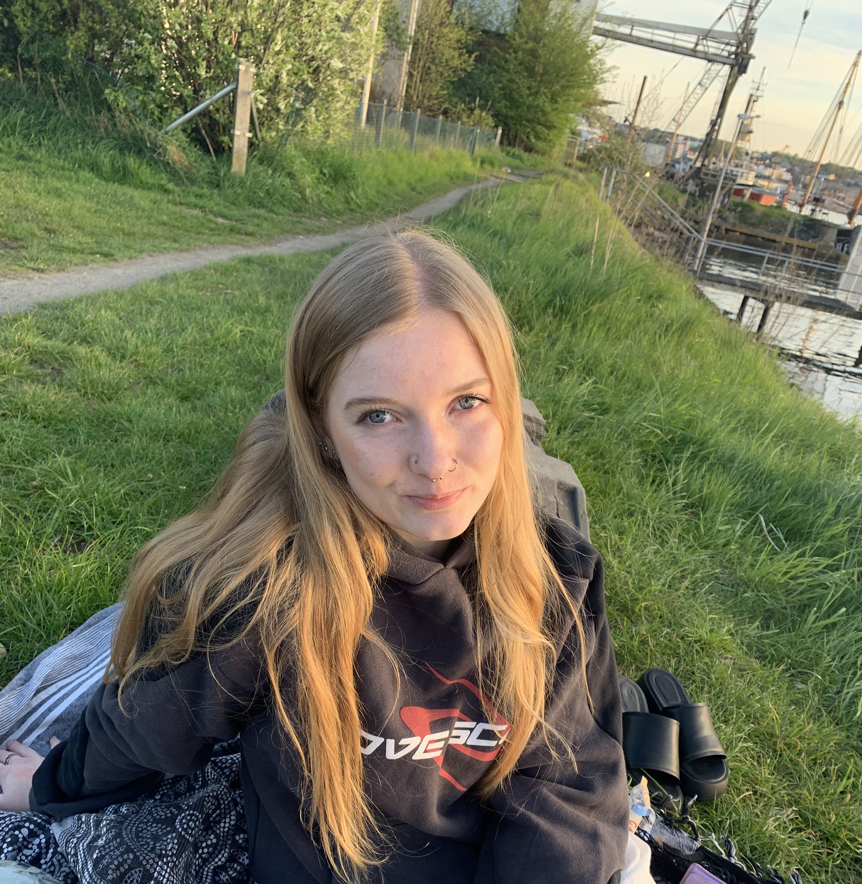 Black and white portrait of a young woman with long blonde hair sitting on a grassy riverbank, wearing a casual hoodie. She looks gently at the camera. In the background are cranes and boats at a harbor.
