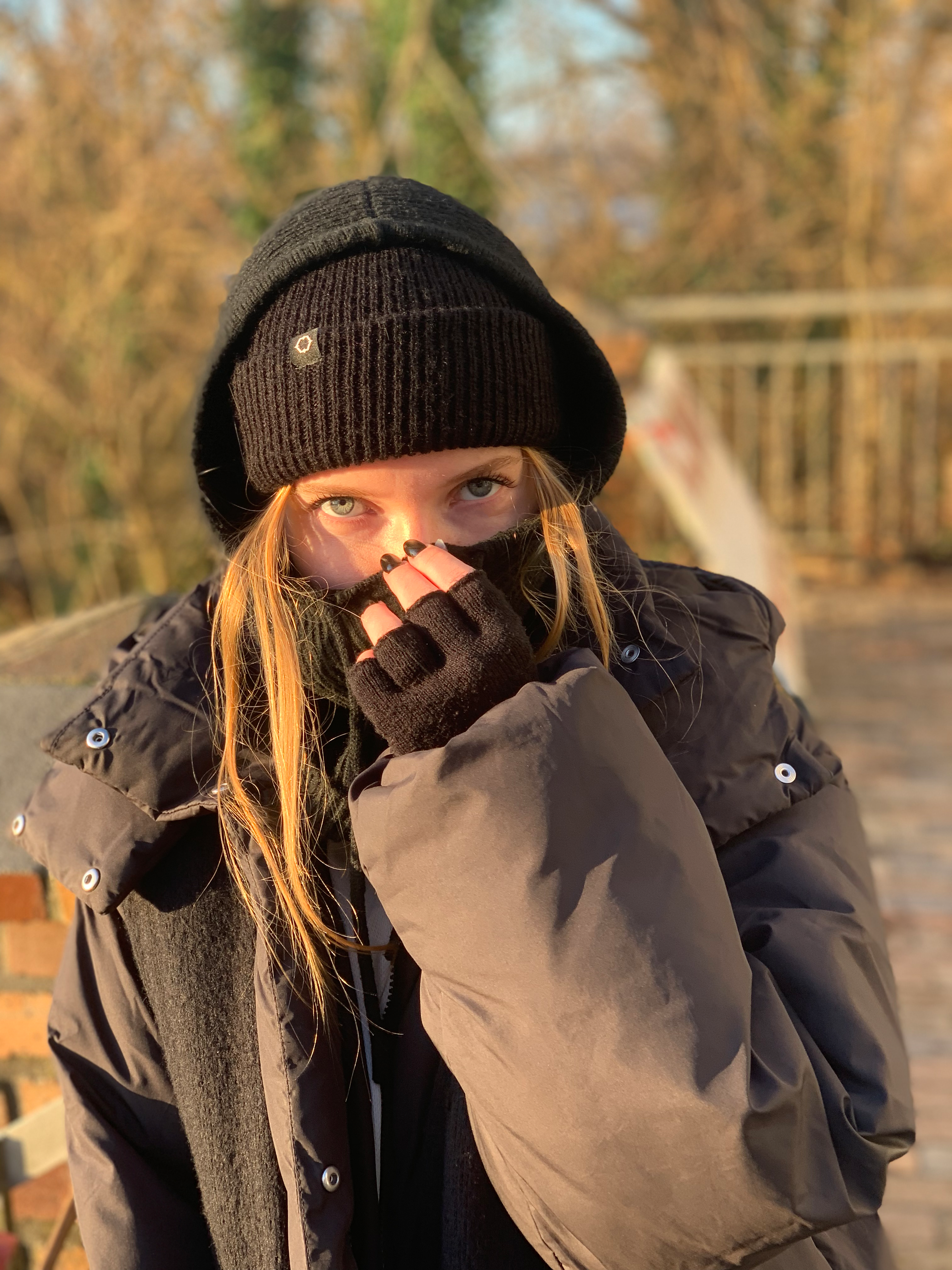 Black and white photograph of a young woman wearing a thick dark jacket, black knit hat, and gloves, standing outside on a bridge. She playfully covers the lower half of her face with her gloved hand. The lighting emphasizes her eyes and facial expression.